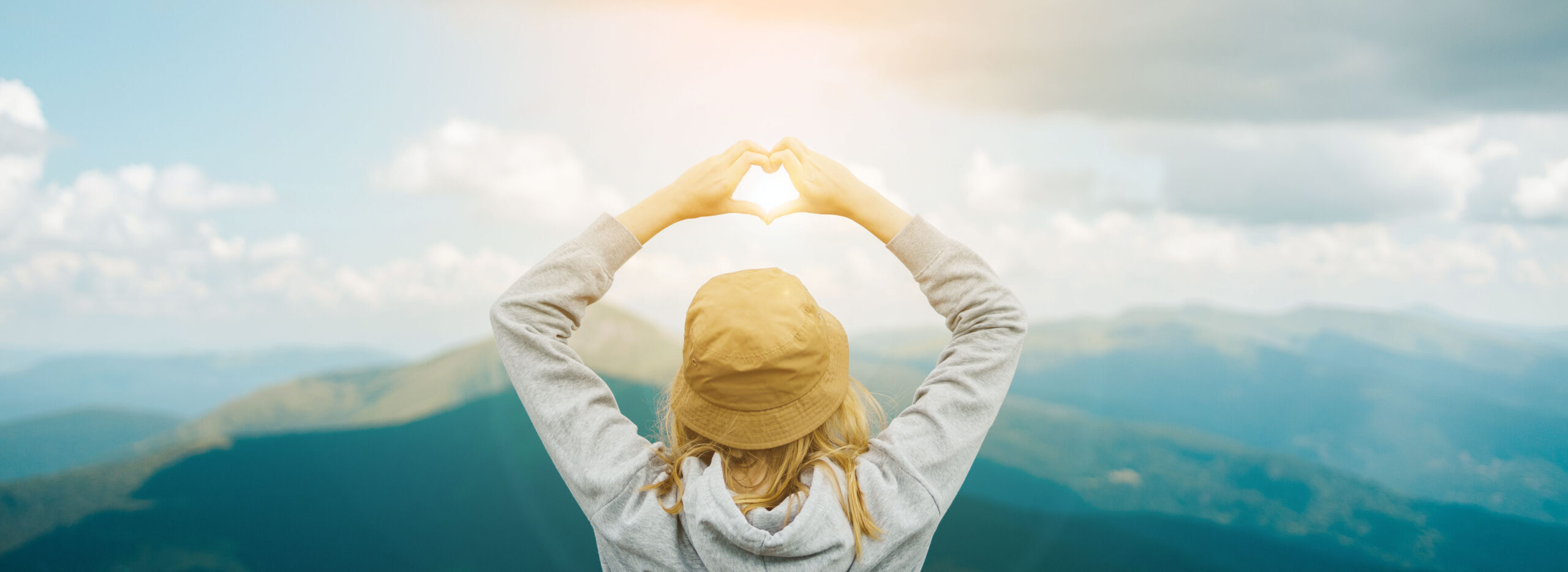 A woman making a heart shape with her hands, standing in the mountains